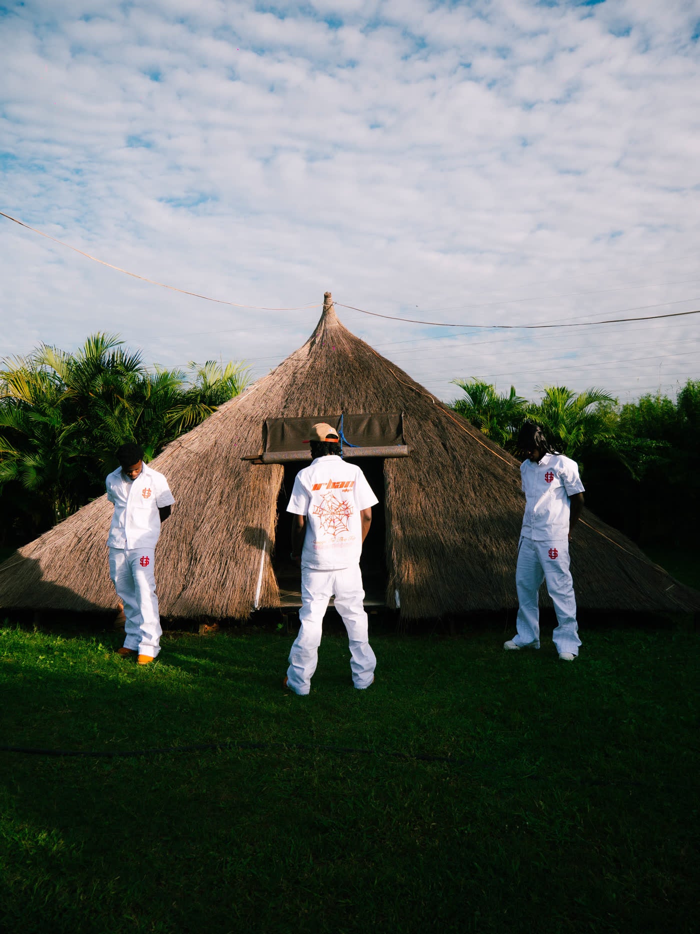Models in white denim walking across grass toward a thatched hut under open sky