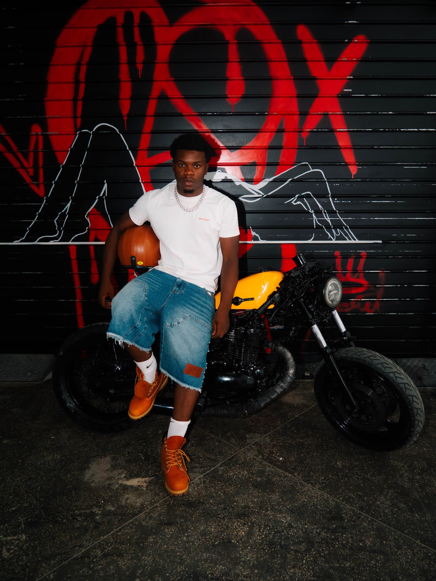 Model seated on a yellow motorcycle in a white tee and denim shorts before a red graffiti shutter